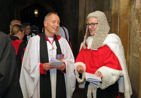 The Venerable Jonathan Chaffey, a priest wearing a white surplice, black preaching scarf and several medals, converses with Mr Justice Murray, a senior high court judge in ceremonial robes and wig, standing in the Cathedral cloisters