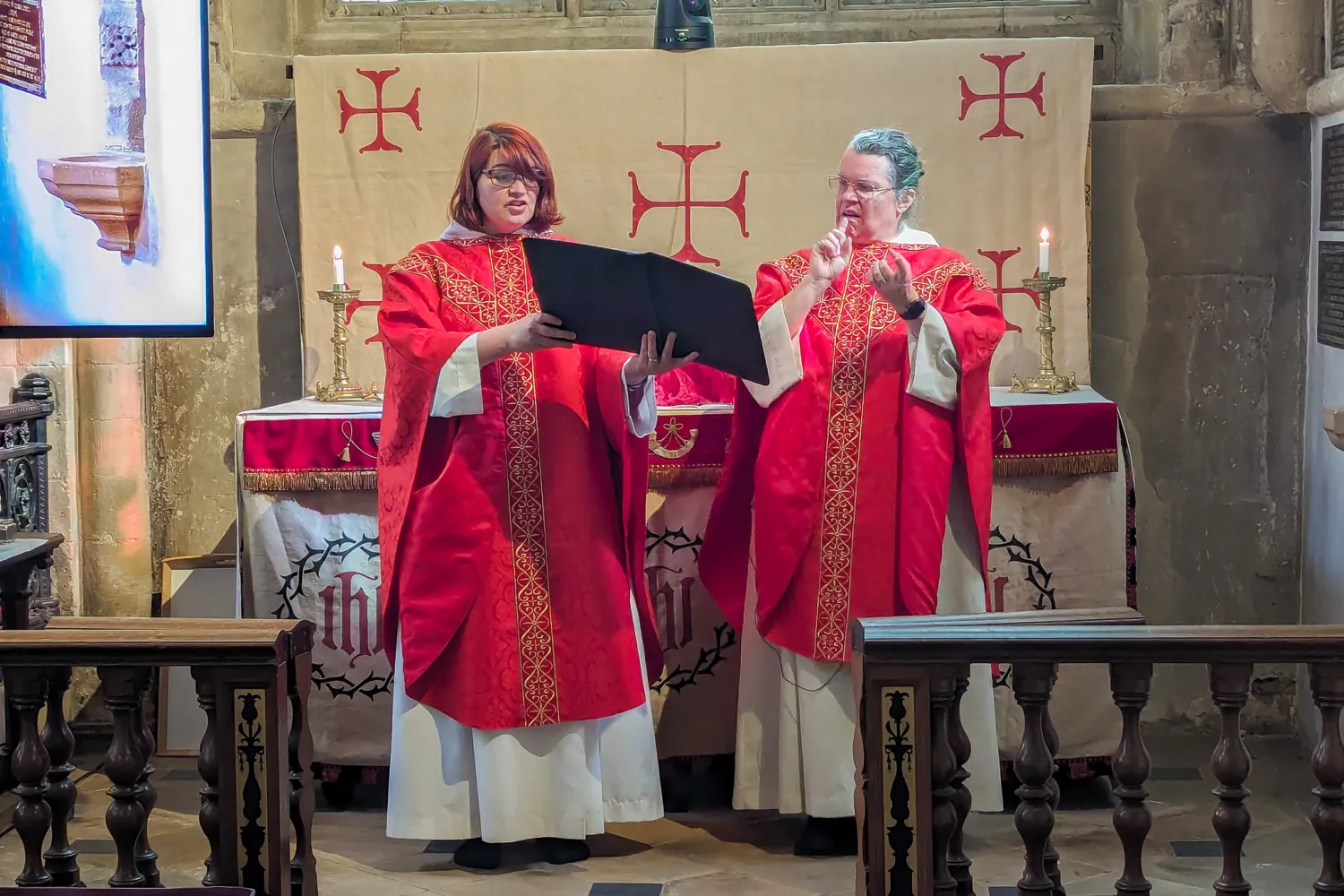 Revd Philippa White and Reverend Hannah Lewis celebrate the eucharist together. Revd Philippa is holding the book of the liturgy, while Revd Hannah is reciting the liturgy in British Sign Language, her hands in mid air. They are both wearing red chausibles, appropriate to the final weeks of Lent, and standing in front of the Remembrance Chapel altar.