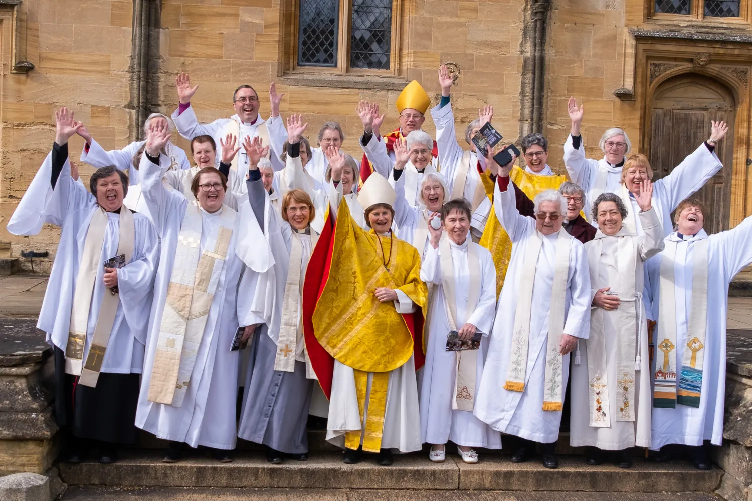 A large group of women ordained in 1994 are gathered wearing albs and stoles, and joined by Bishops Olivia Graham, Steven Croft and Gavin Collins. The first female Dean of Christ Church, Sarah Foot, is also present. They are waving and looking thrilled that women triumphed over discrimination to flourish in spirit and ministry.